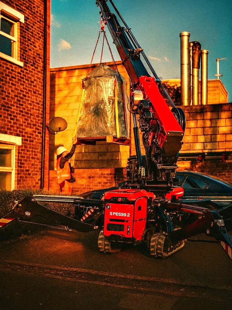 Red spider crane lifting a wrapped crate beside a brick building at dusk